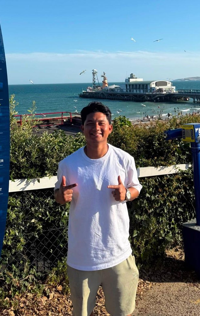 Nikhil smiling on a sunny clifftop above a pier and the sea